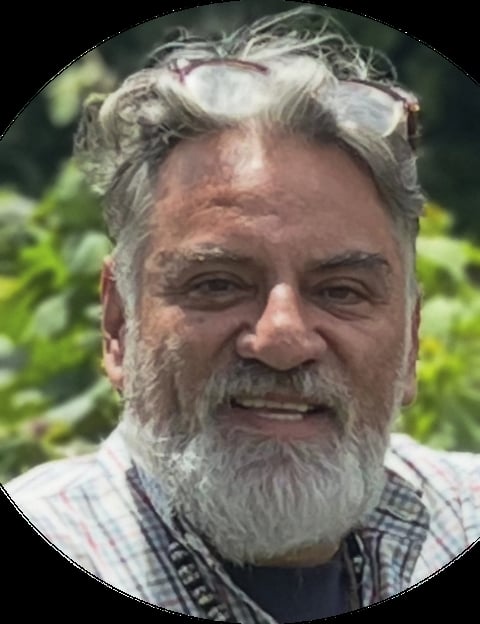 Portrait of an older man with gray hair and full beard, wearing a plaid shirt and sunglasses on his head, smiling outdoors