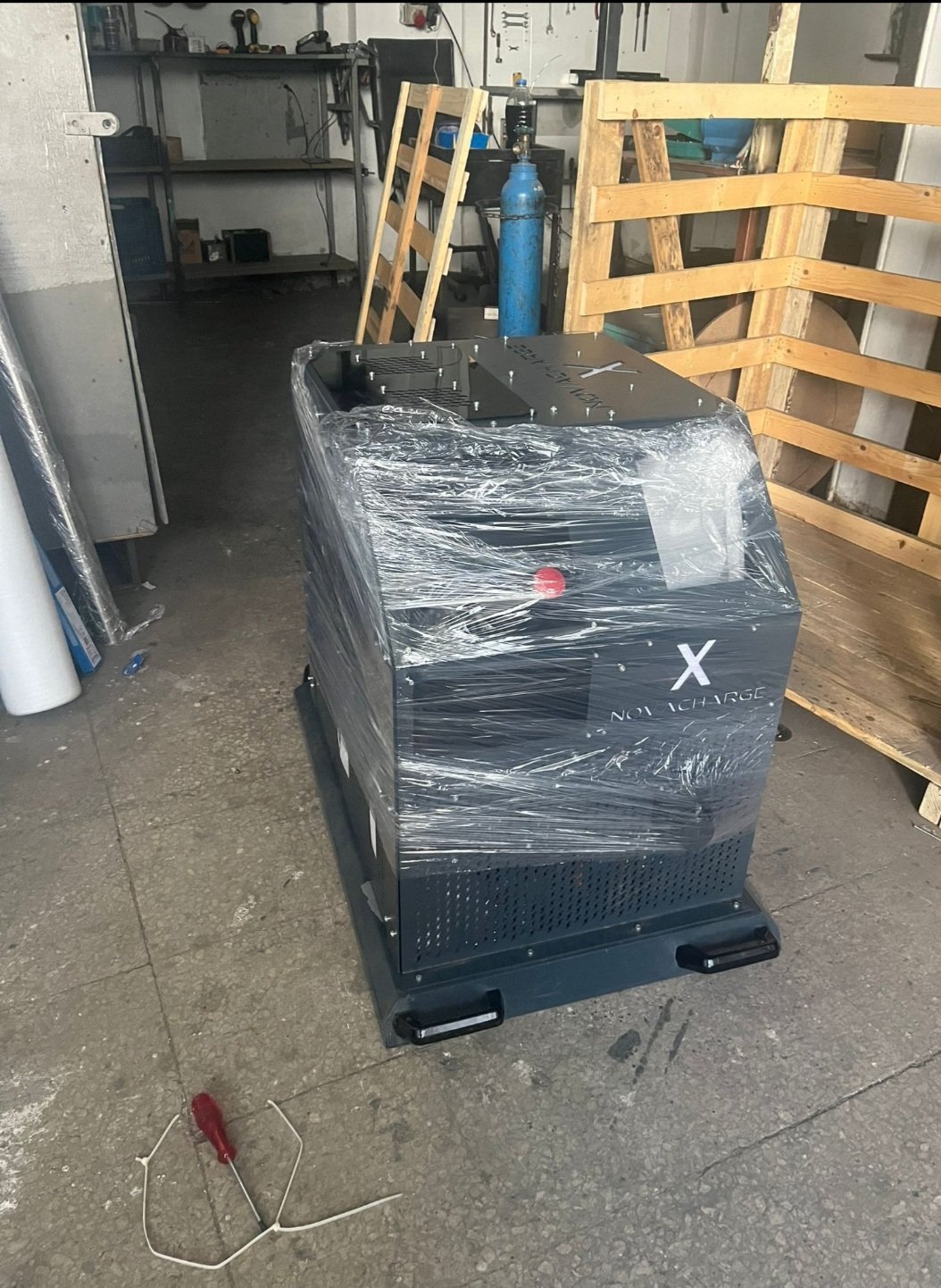 Black industrial equipment wrapped in plastic inside a warehouse or storage facility with wooden pallets and shelving visible