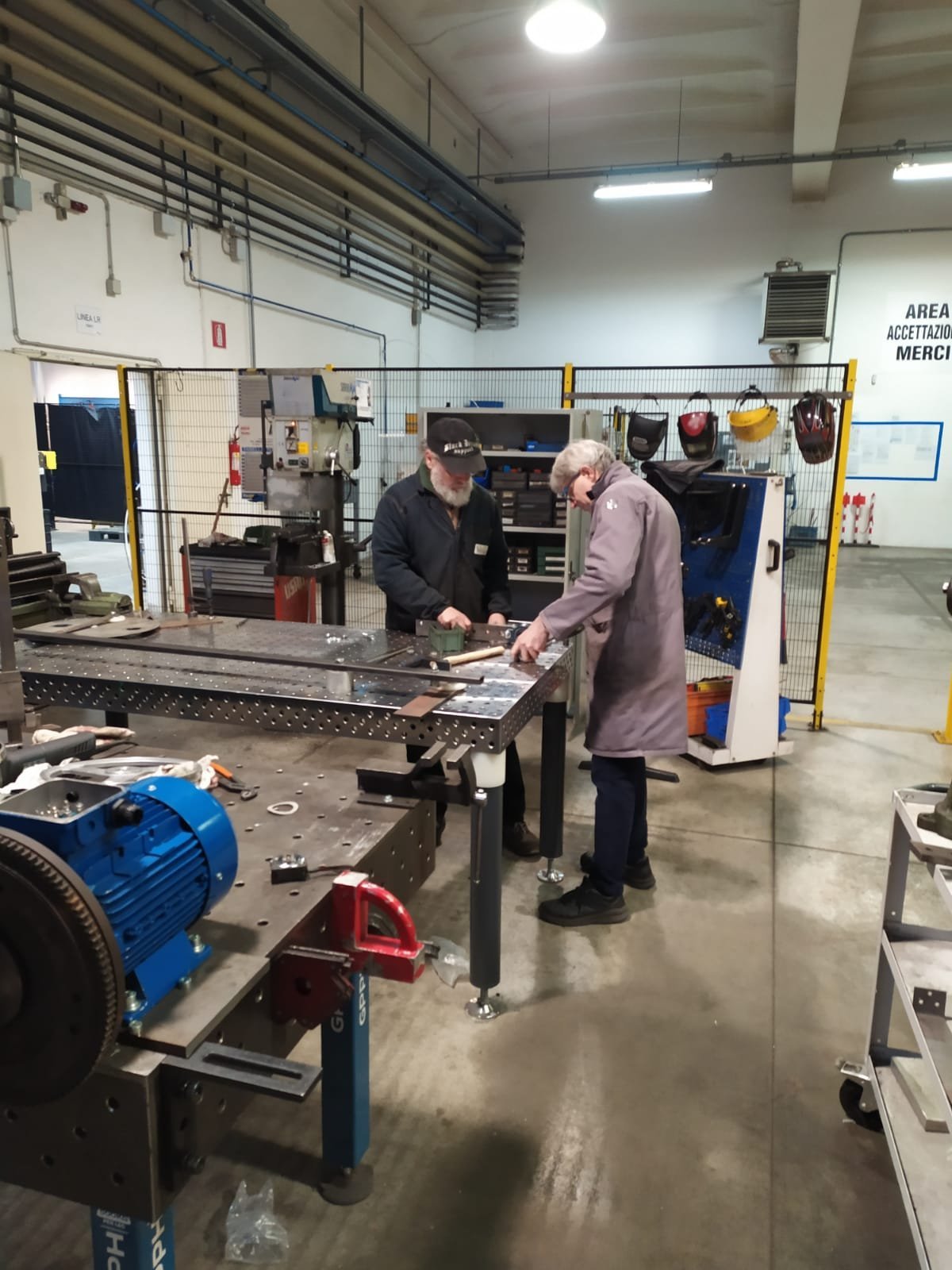Two workers in an industrial workshop examining metal parts on a workbench surrounded by machinery and tools